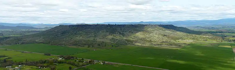 A flat topped mesa surrouned by cliffs and tree-covered slopes with farmland below